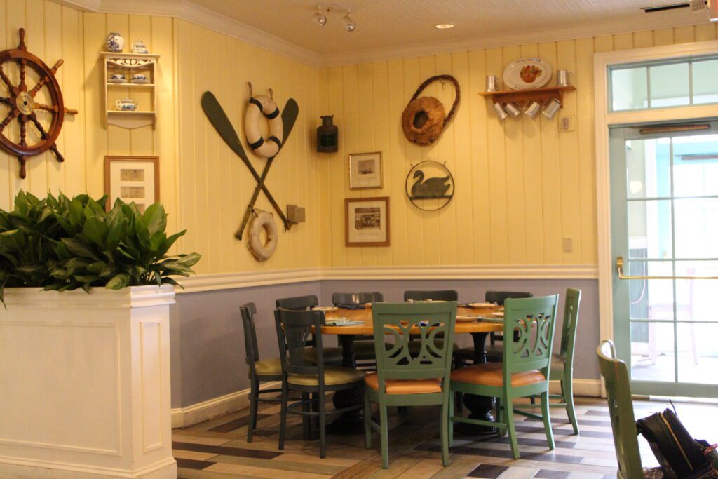 Olivia's Cafe is the main and only real non quick service option of the Disney's Old Key West Resort restaurants. Inside it has a coastal, vintage theme with bright walls featuring nautical items like paddles and a lifesaver. A large round table is pictured in a corner next to greenery.