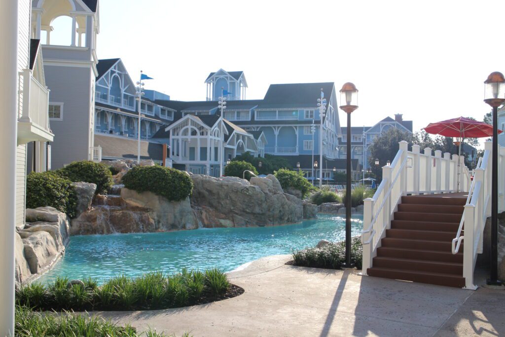 Stormalong Bay pool at Disney's Beach Club Resort sparkles light blue in the sun surrounded by rocks, landscaping and stairs to a bridge over the water