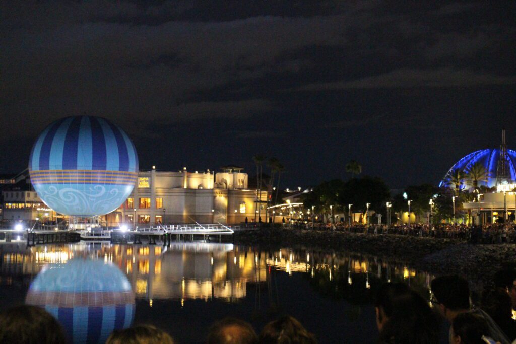 A nighttime view of the water's edge at Disney Springs with the balloon attraction and crowds facing the water.