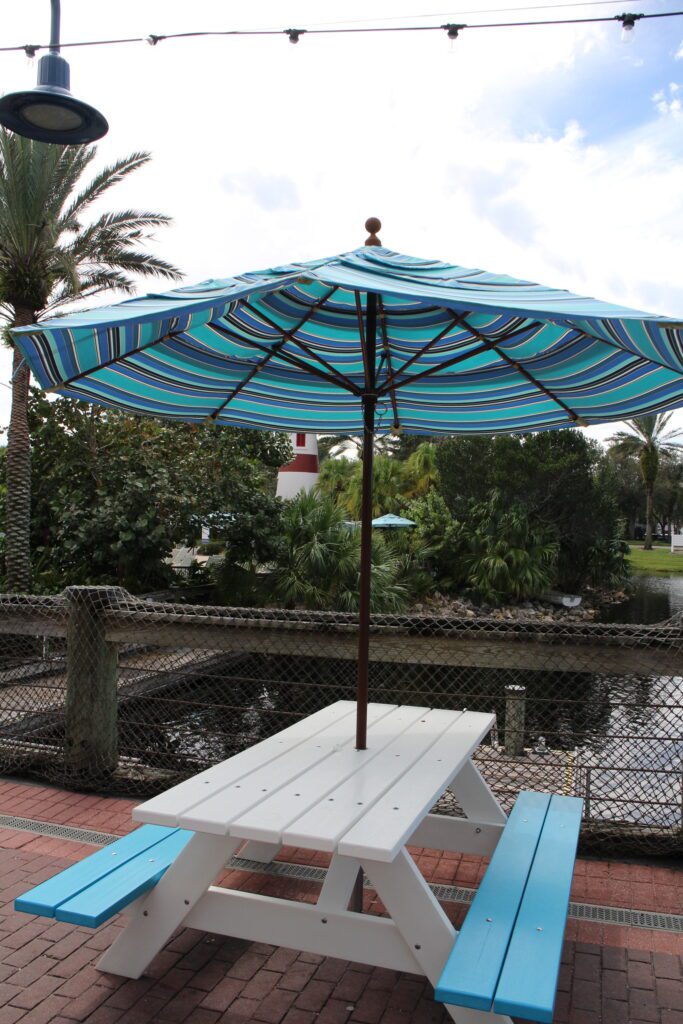 A white picnic table with blue bench seating, covered by a blue striped umbrella next to the water way at Disney's Old Key West Resort.