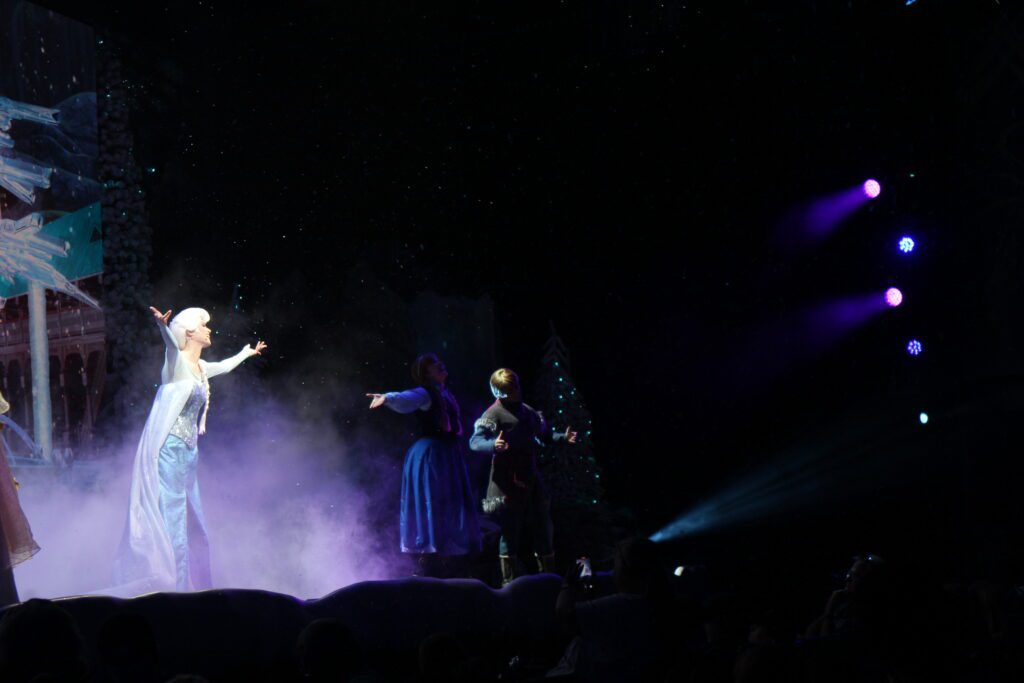 Elsa stands on a smoky dark stage in her iconic icy blue gown with her arms open to the audience at the singalong at Hollywood Studios. This is the only show of Frozen at Disney World.