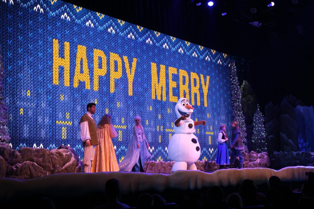 The Arendelle historians, Elsa, Olaf, Anna and Kristoff stand on stage at the Frozen singalong in front of a holiday sweater inspired screen that says "Happy Merry."