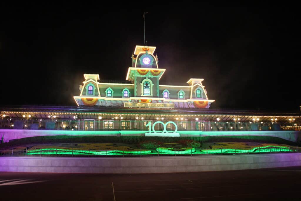 Magic Kingdom entrance at night with the train station decorated for fall with orange bunting and lit in green and purple.