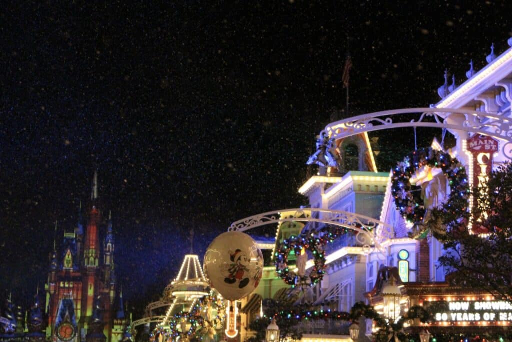 Snow falls on Main Street USA at Magic Kingdom at night, with the buildings lit up, during the Disney World Christmas season.