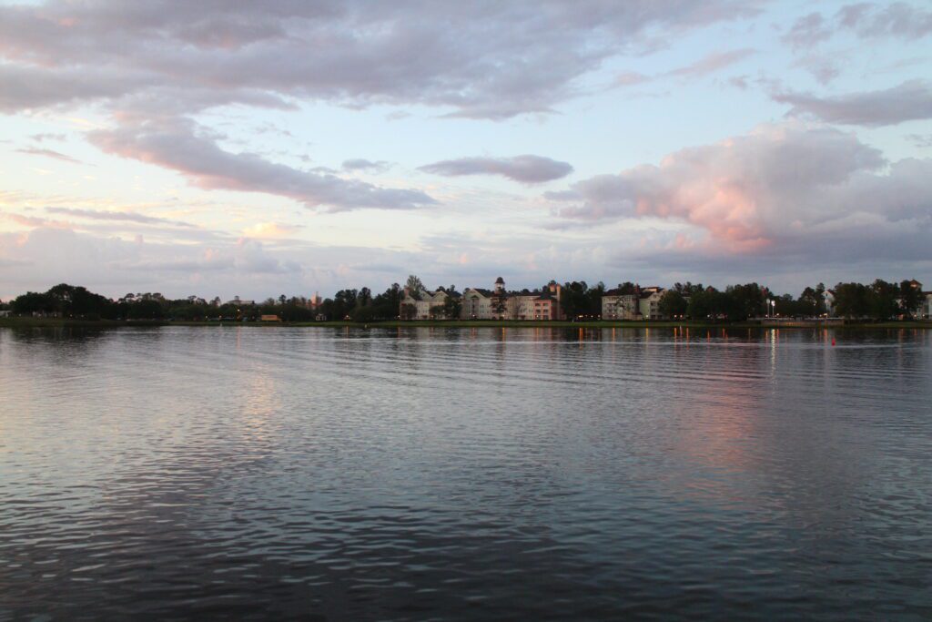 Views of the water at Disney Springs during sunset. The sky is pastel and off in the distance are brightly colored Saratoga Springs resort buildings.