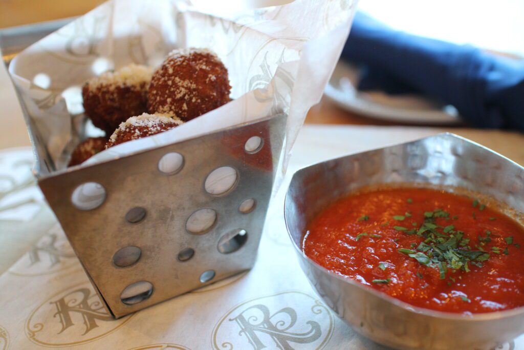 A silver basket of crispy, brown arancini balls topped with powdered cheese next to a bowl of red marinara sauce with a green herb on it. This is from Bar Riva at Disney's Riviera Resort which has some of the best dining of the DVC home resort options.