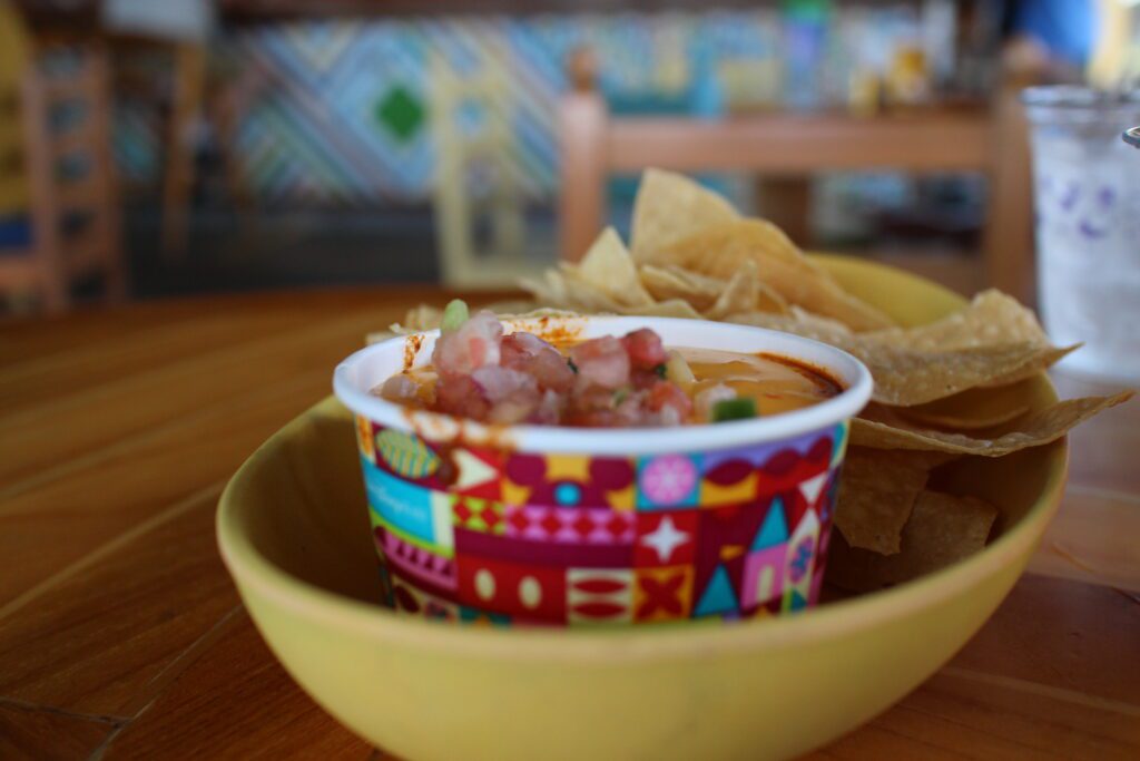 Banana Cabana Caribbean Chips and Dip in a yellow bowl with a cheesy dip covered in tomatoes. There are lots of different bar and lounge style foods to try on your Disney Skyliner bar crawl.