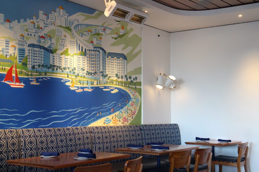 A Mediterranean style, colorful wall mural with lots of blues shows the Riviera resort on the water in a corner of the restaurant where there is hightop booth seating.