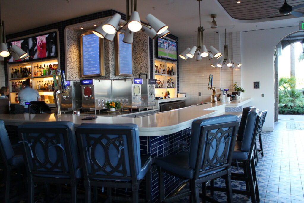 The Bar at Bar Riva with blue stools with backs, white counter top, tiled wall with a menu and televisions.