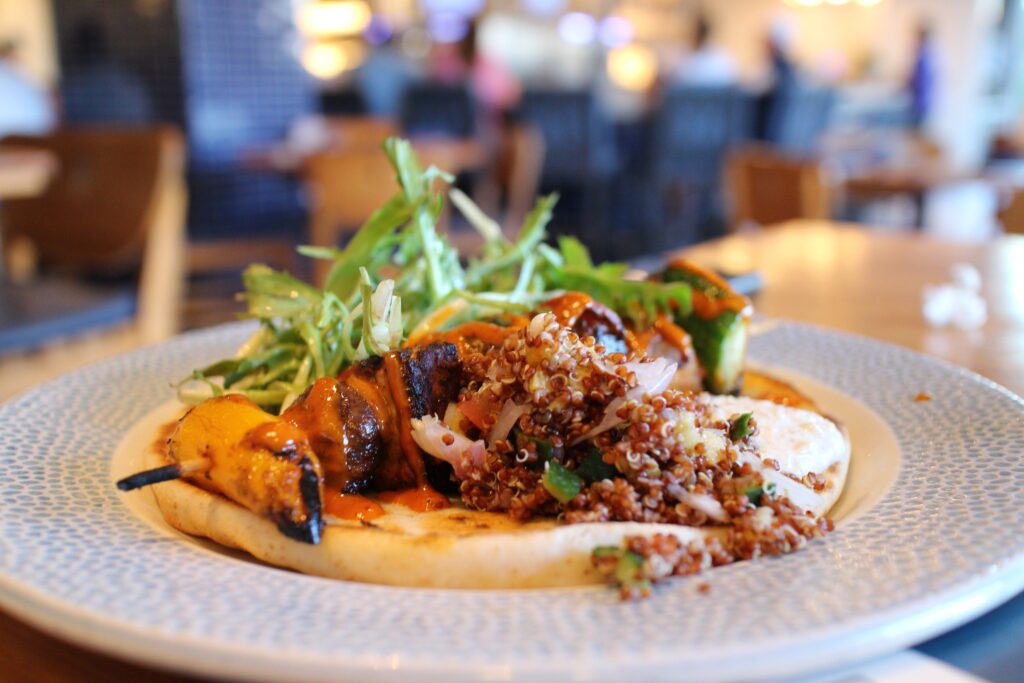 The plant based Bar Riva Grilled Vegetable Skewer with Harissa-marinated Vegetable Skewer, Plant-based Cucumber Yogurt, Tomato Lemon Vinaigrette and Quinoa Salad served over a Grilled Pita.