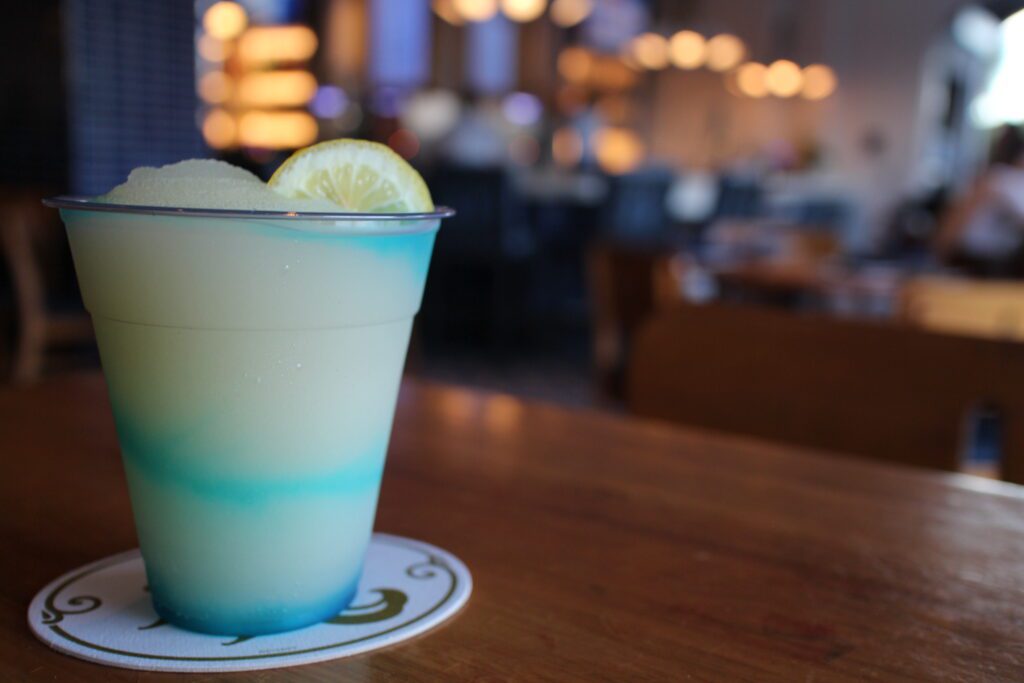 The Mediterranean Wave drink at Bar Riva is a light green frozen, icy style drink with blue swirls in a clear cup, sitting on a brown table with the out of focus chic Disney's Riviera Resort bar and lights in the background.