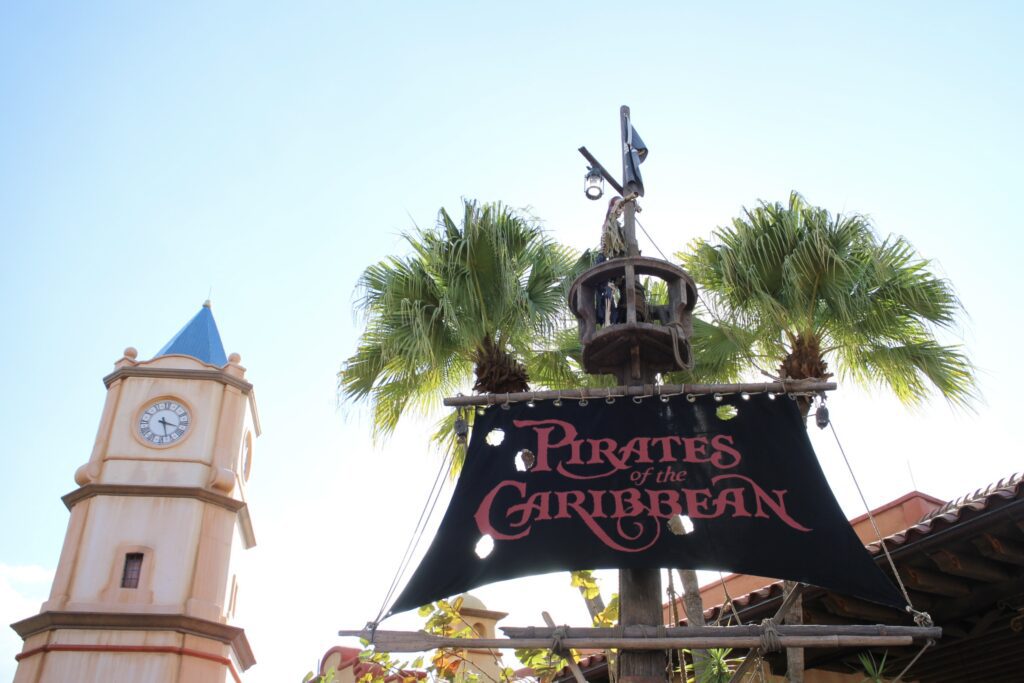 The Pirates of the Caribbean attraction sign, on a black ship sail with letter in red writing. A clock tower in a vintage architectural style sits on the left. A palm tree is behind the sail sign.