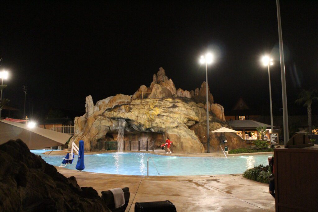 The Polynesian pool at night with crystal blue water, a large volcano structure and a Disney World pool bar off to the right.