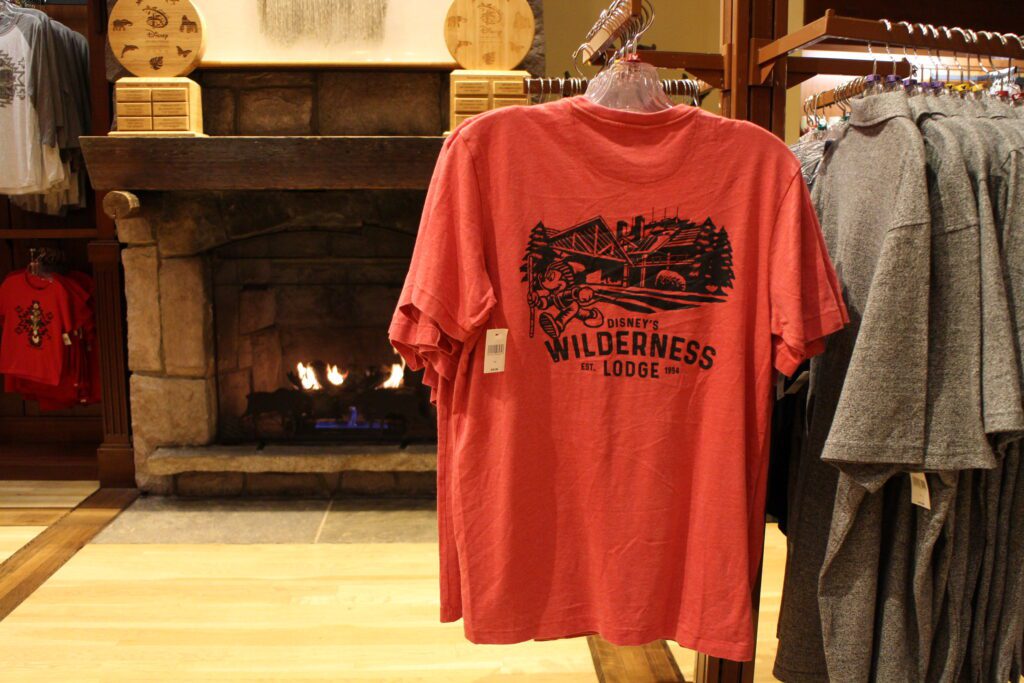 A red Wilderness Lodge shirt with the resort and Mickey on it in black ink in front of a cozy fireplace in the themed gift shop.