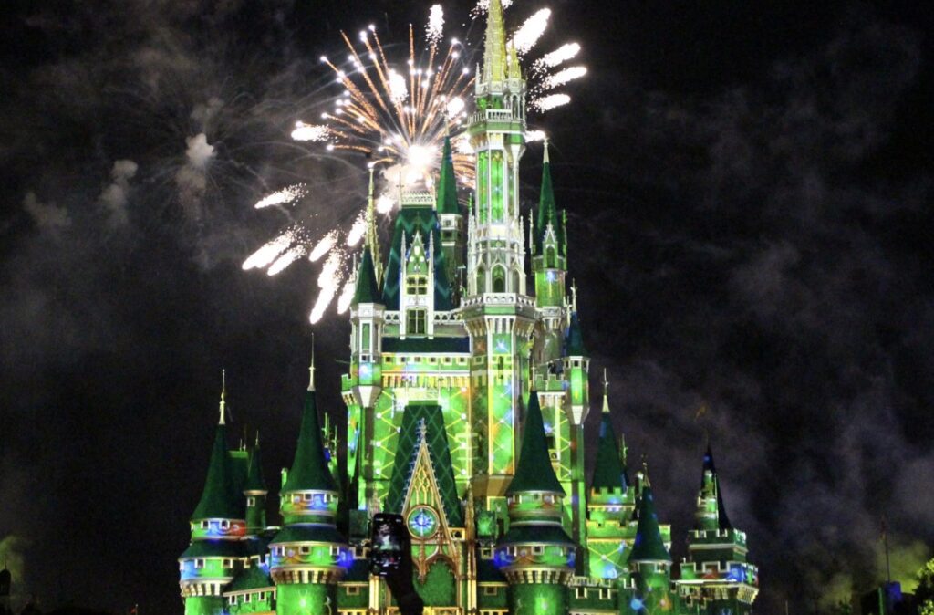 The Disney castle lit green like a Christmas tree with fireworks over it in front of a dark night sky.