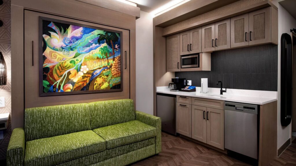 The Polynesian Island Tower Deluxe Studio concept image showing a pull down bed behind a couch and a larger kitchenette with a dishwasher.