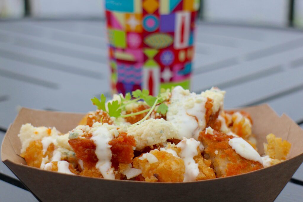 A poutine style dish sits on a table in a paper basket, garnished with greenery and a colorful Disney World drink cup in the background.